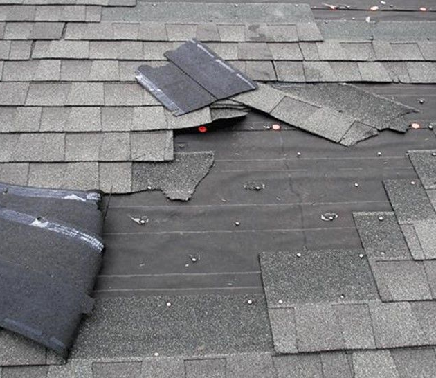 Damaged asphalt roof shingles missing and peeled back, exposing the underlying black roofing felt and nails.