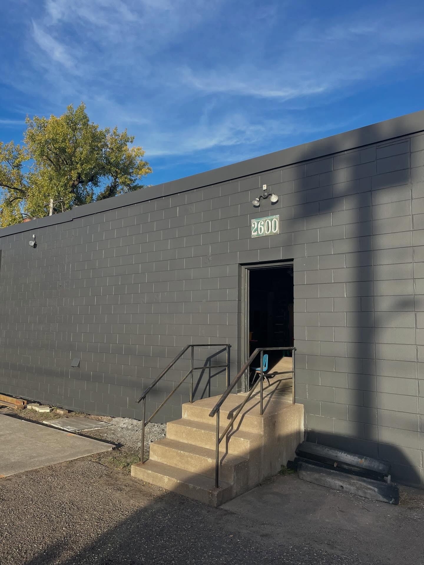 A dark gray cinder block building with a doorway, stone stairs, and a metal railing under a blue sky.
