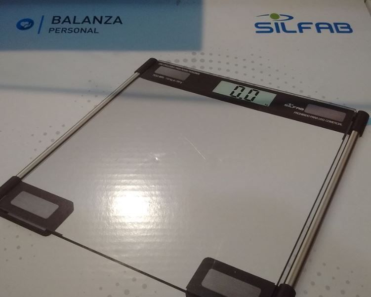 Balanza digital para baño con marco negro y plataforma blanca sobre una caja de producto con logotipos.