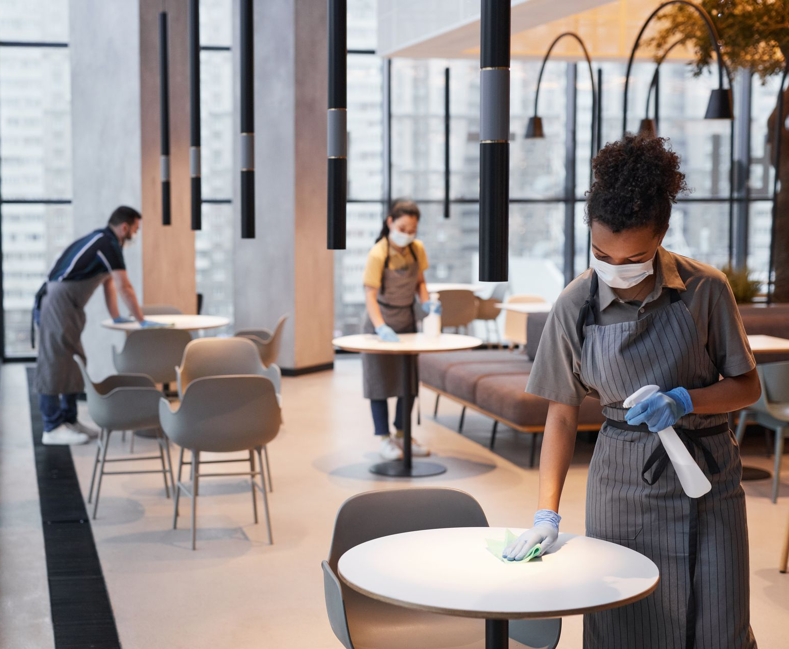 Staff wearing masks disinfect tables in a modern restaurant lobby.