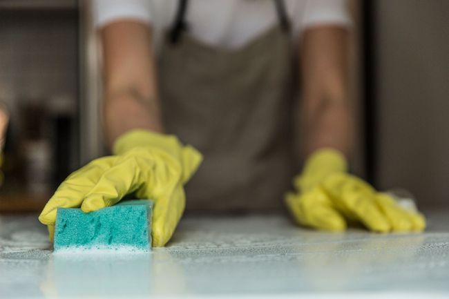 Yellow-gloved hands wiping a countertop with a blue sponge in a blurred kitchen setting