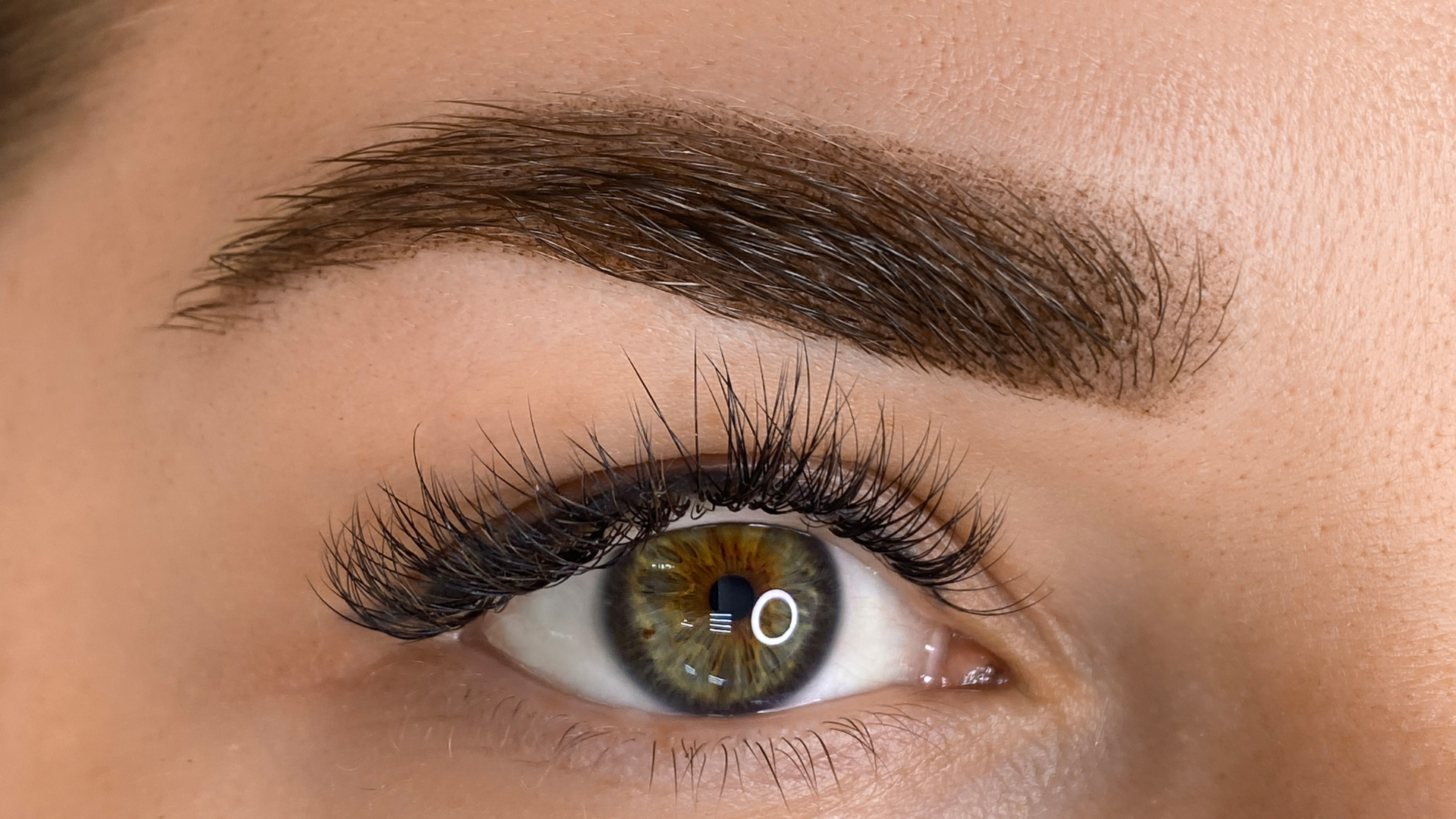 Close-up of a person's eye with thick, dark eyelashes, a defined eyebrow, and a green iris with a light ring.