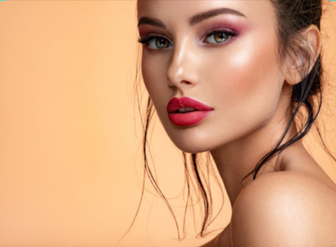Woman with pink eye shadow and red lipstick, looking towards the camera, against a peach background.