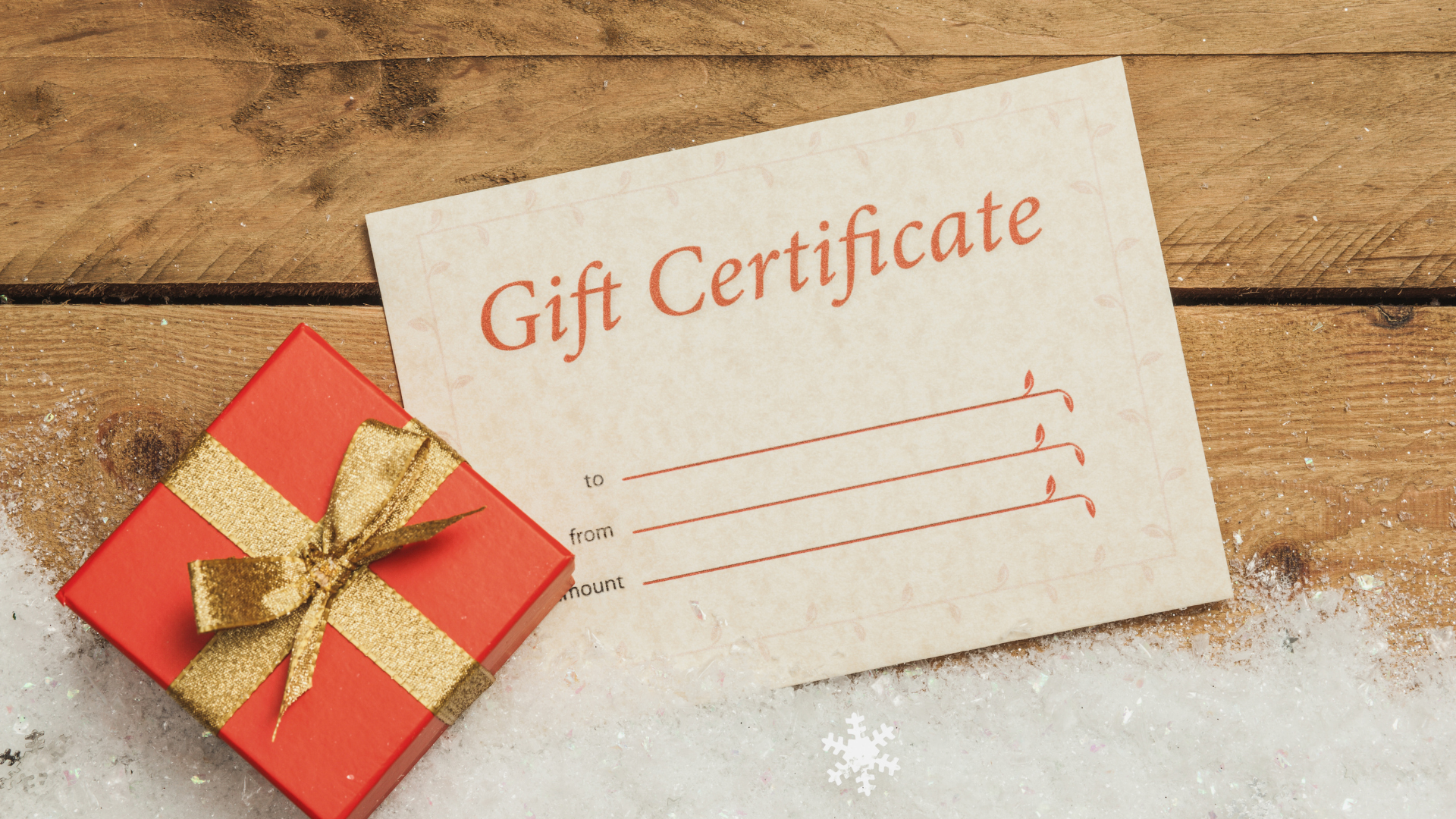 Red gift box with gold bow and gift certificate on a wood surface covered in snow.