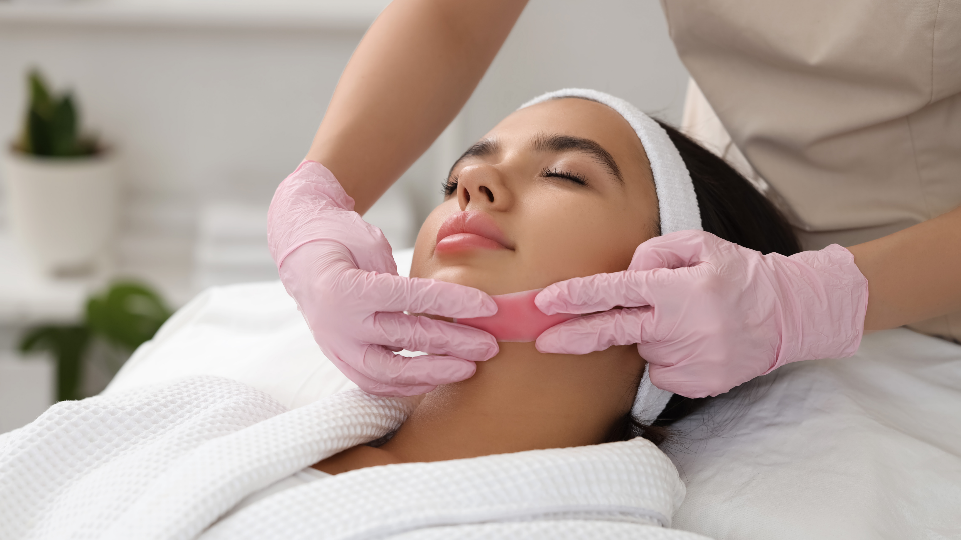 Woman receiving facial wax treatment in a spa, wearing a robe. Hands in pink gloves applying wax strip.
