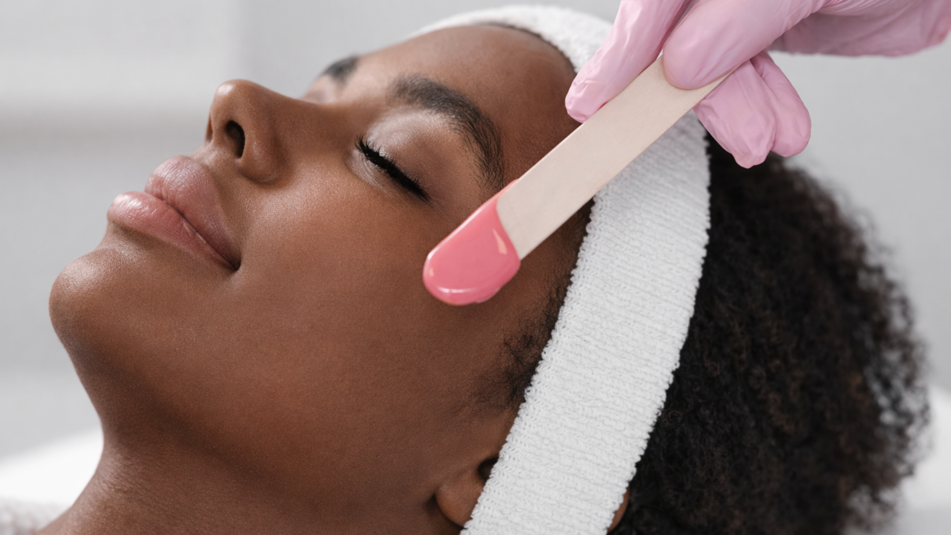 Facial waxing: A person lies down while a gloved hand applies wax to the cheek with a wooden applicator.