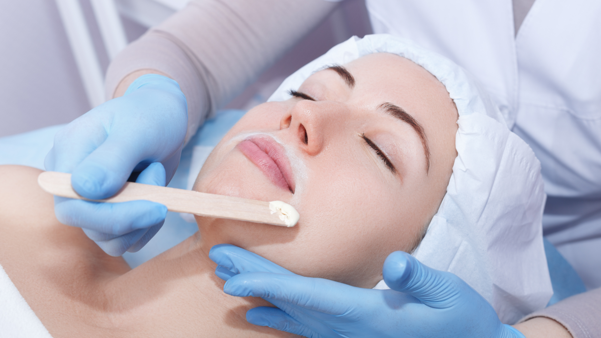 A woman's face prepped for waxing, the esthetician applies wax to the upper lip with a wooden spatula.