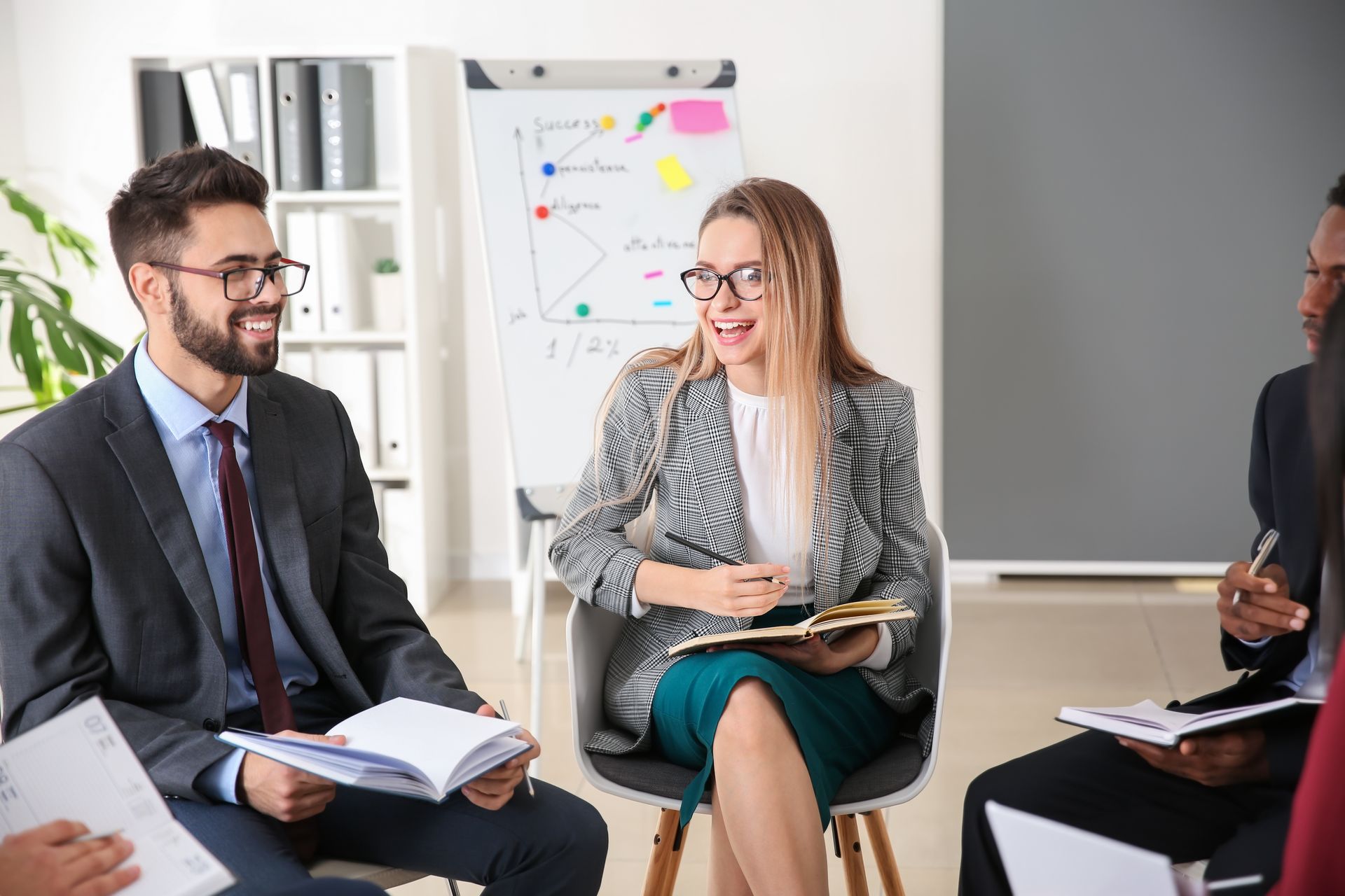 People in business attire seated in a circle, engaged in a discussion; whiteboard and office setting in the background.