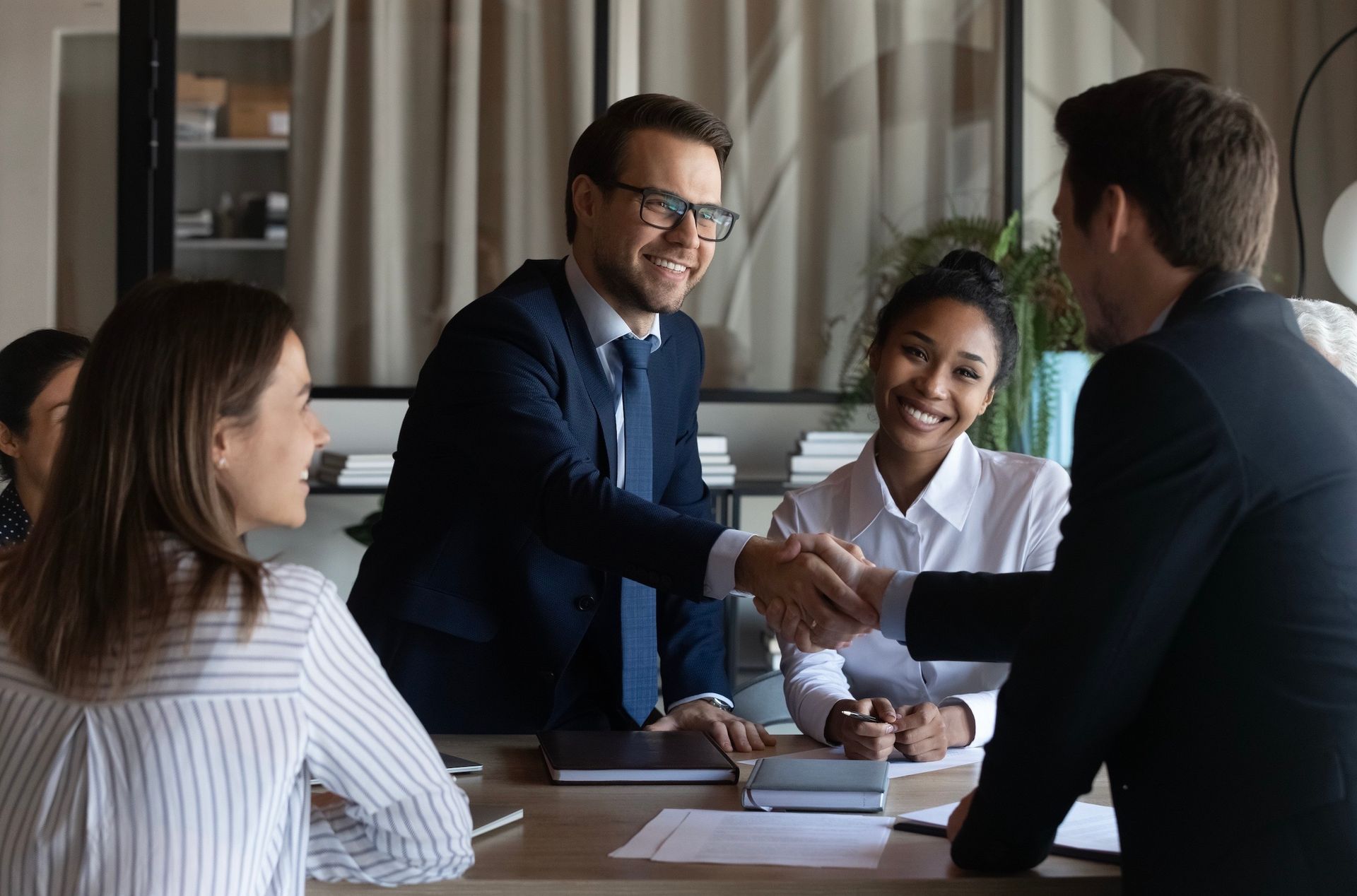 Business meeting: man in suit shakes hands, smiles, others at table.