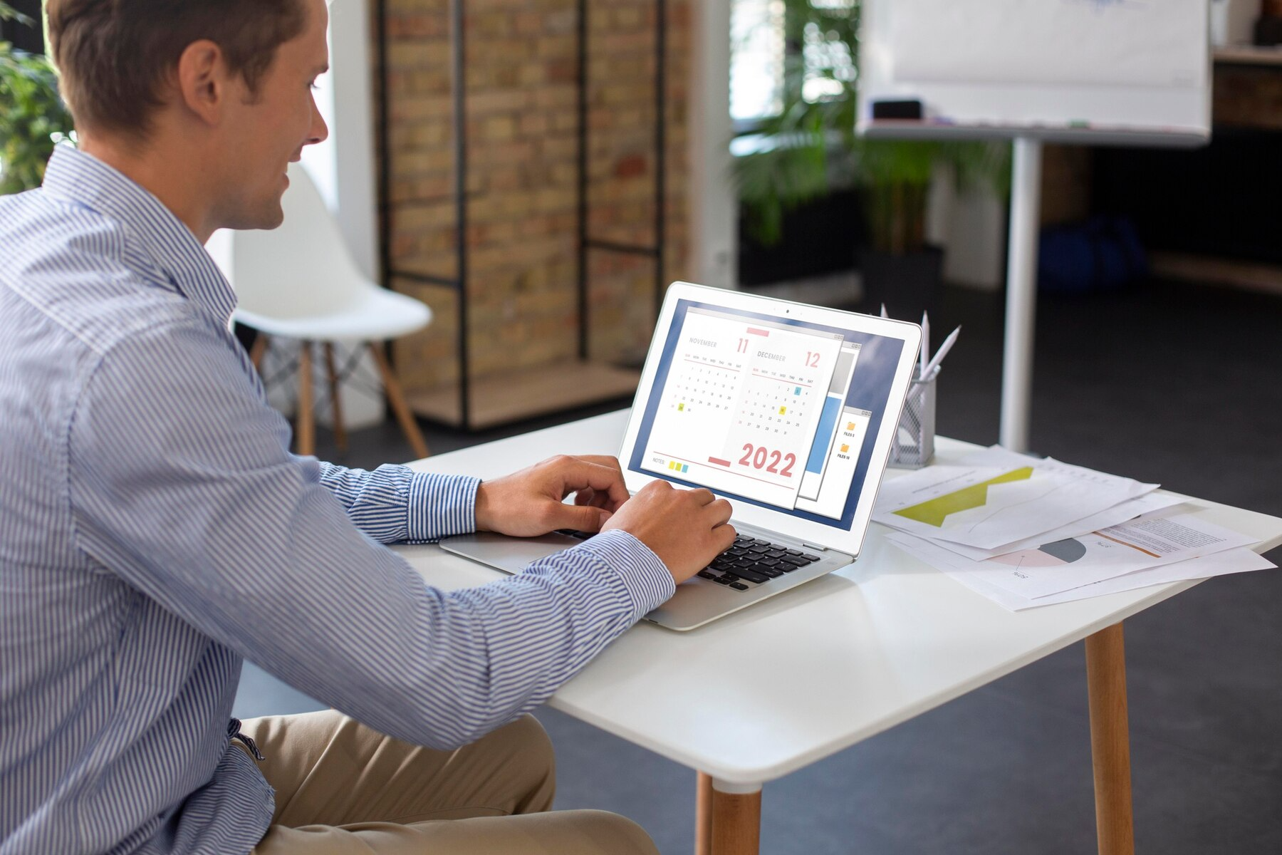 Man in blue shirt working on laptop at a white desk; papers and a cup nearby in a modern office.