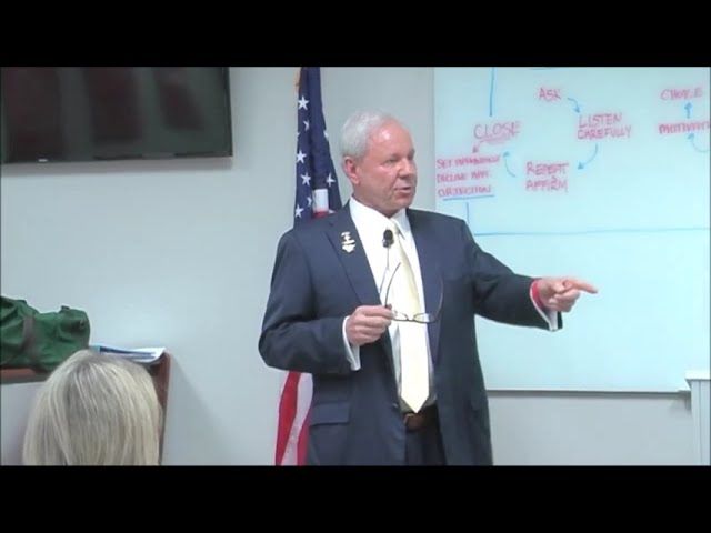 Man in suit gesturing with glasses, speaking in front of a whiteboard with text and American flag.