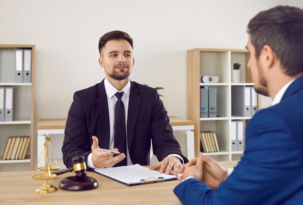 A Man In A Suit And Tie Is Sitting At A Desk Talking To Another Man — Charlestown Law Firm In Central Coast, NSW