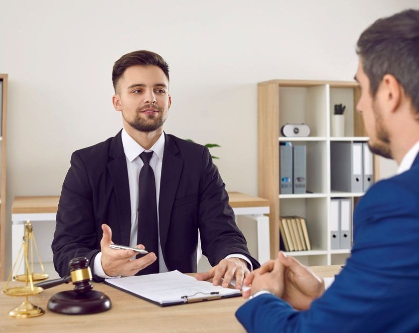 A Man In A Suit And Tie Is Sitting At A Desk Talking To Another Man — Charlestown Law Firm In Lake Macquarie, NSW