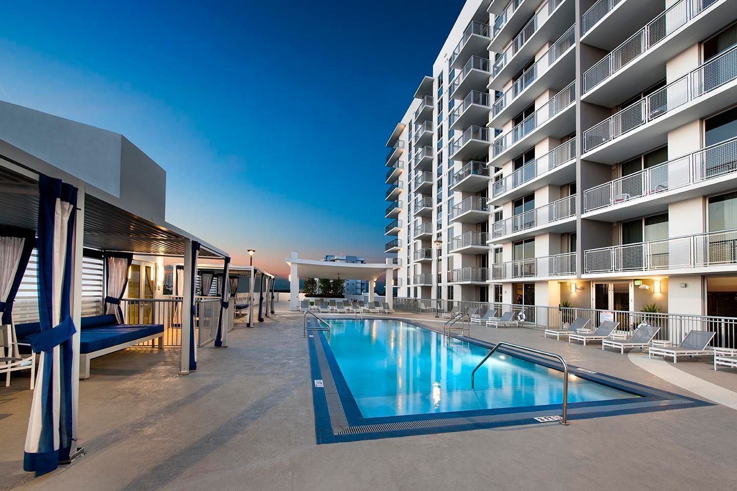 Rooftop pool next to a high-rise building with lounge areas and blue sky.