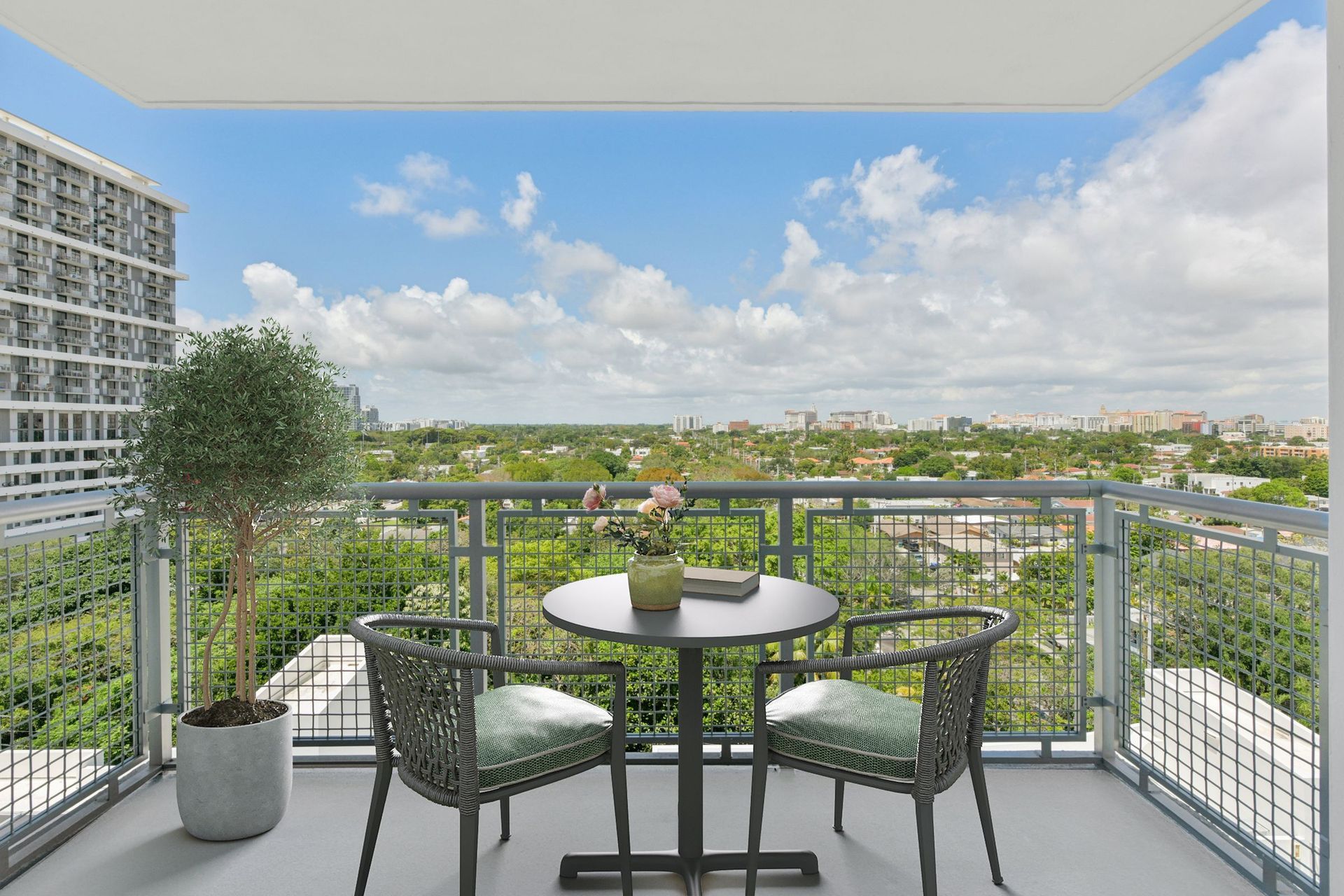 Balcony with two chairs, round table, and potted plant overlooking a city and green landscape.