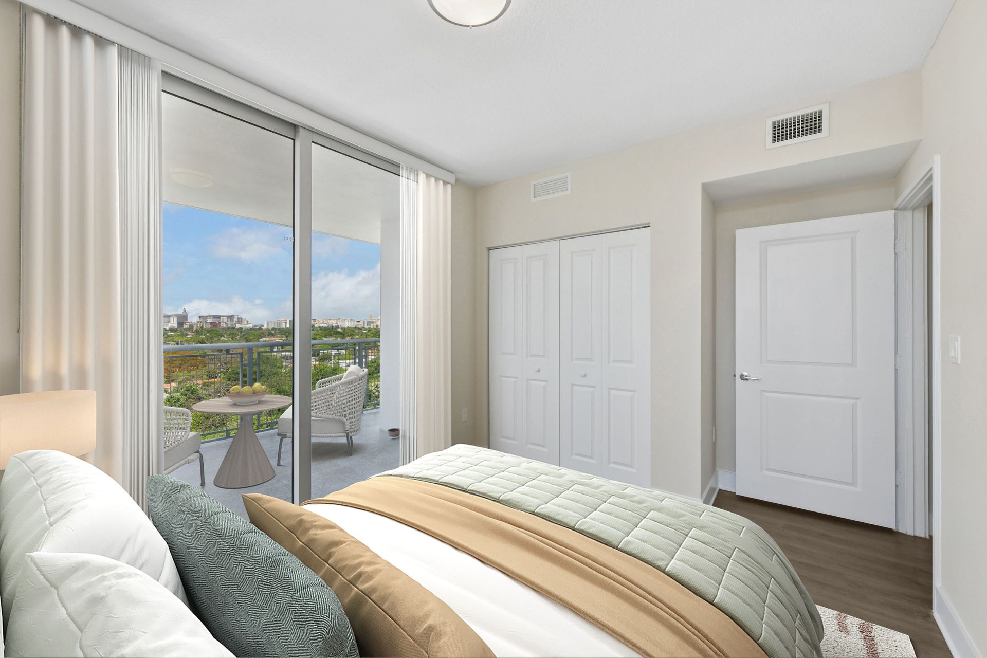 Bright bedroom with bed, large sliding glass doors, and a balcony view of rooftops and sky.
