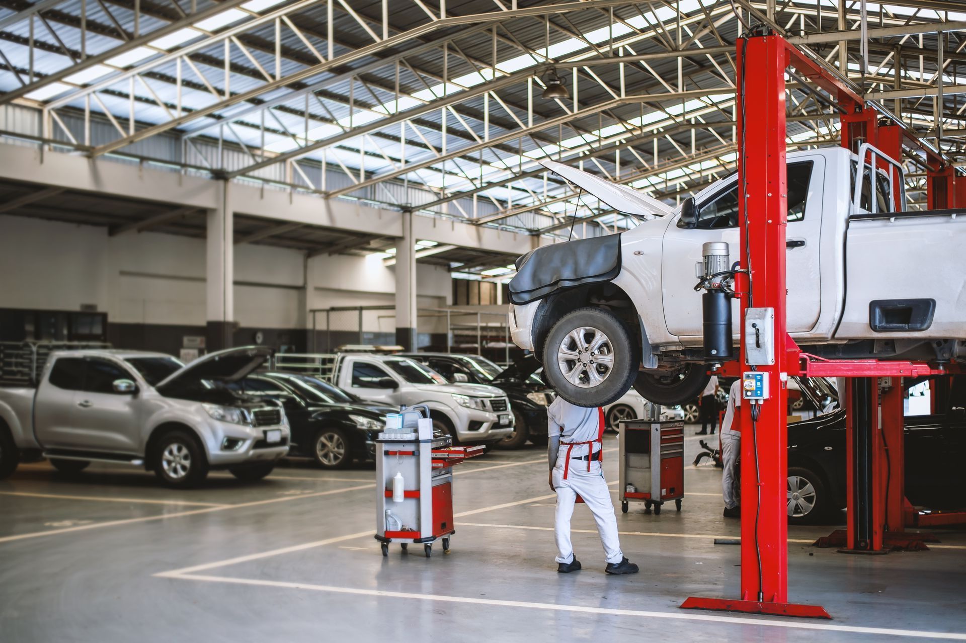 Car repair shop with a lifted white SUV, mechanic underneath, and parked cars in the background