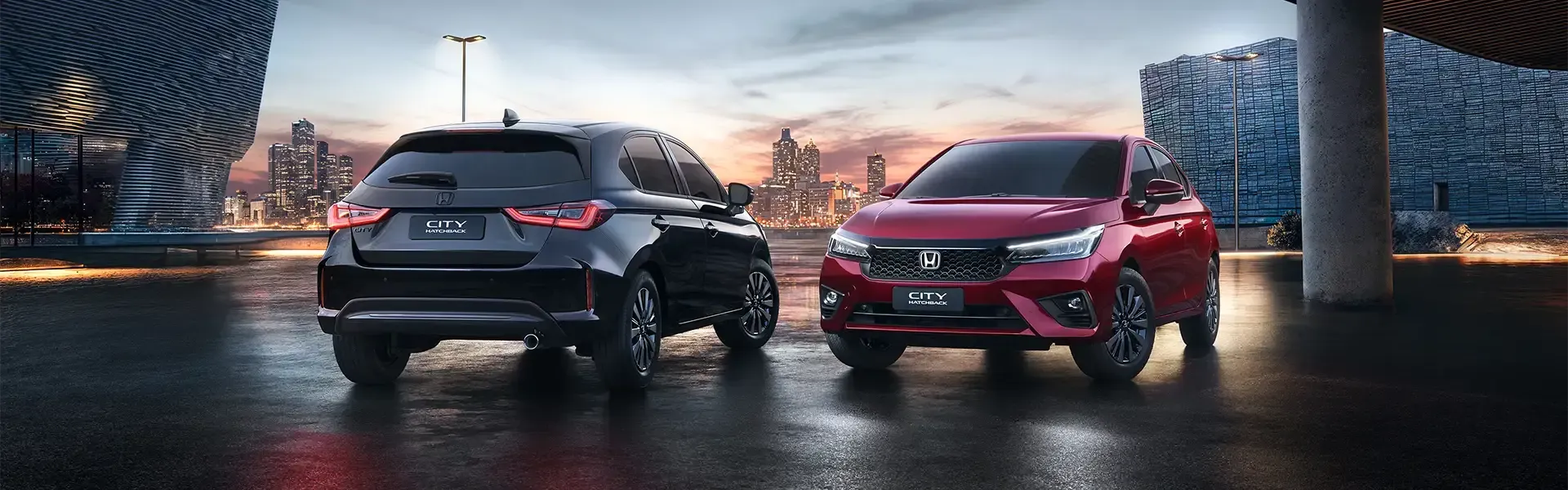 Two parked cars, one black and one red, on a wet street at dusk with modern buildings in the background