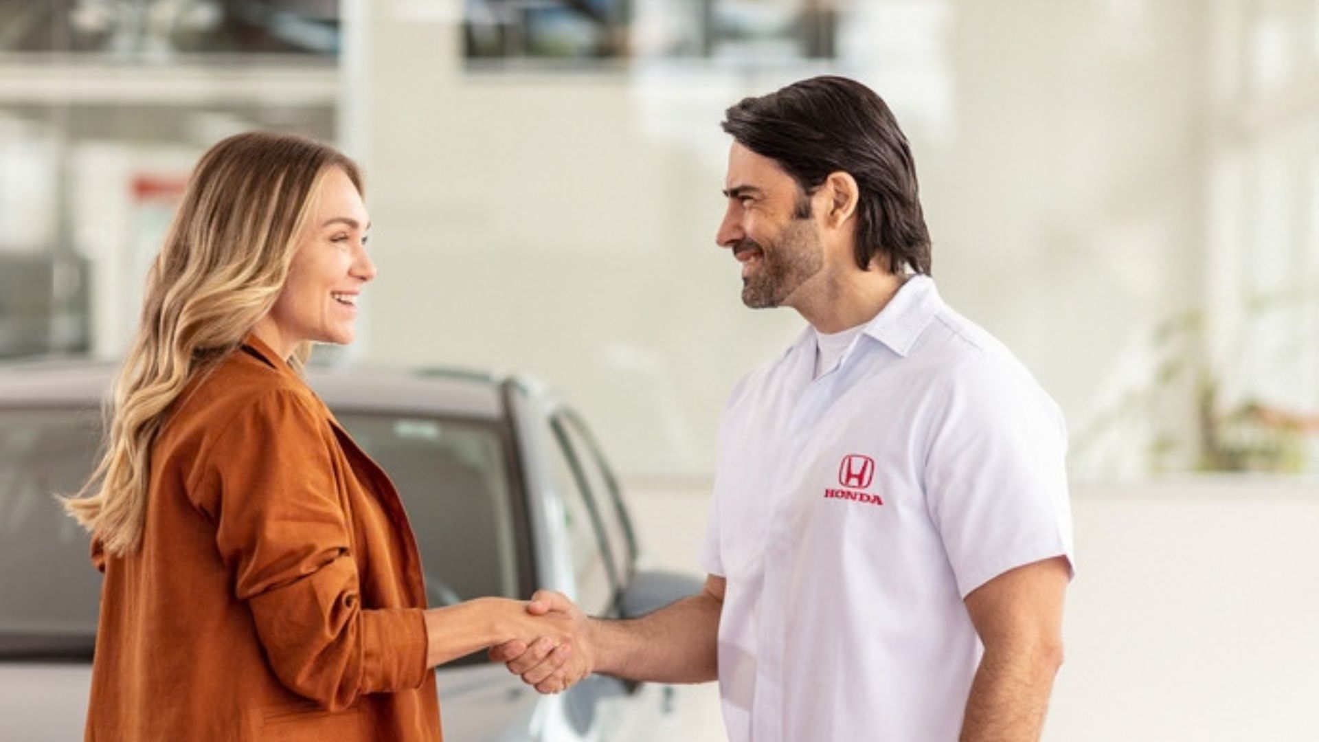 Two people shaking hands in a car showroom, smiling beside a vehicle.