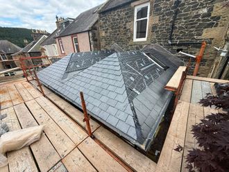 New grey slate roof under construction beside a stone building, with scaffolding and plywood decking