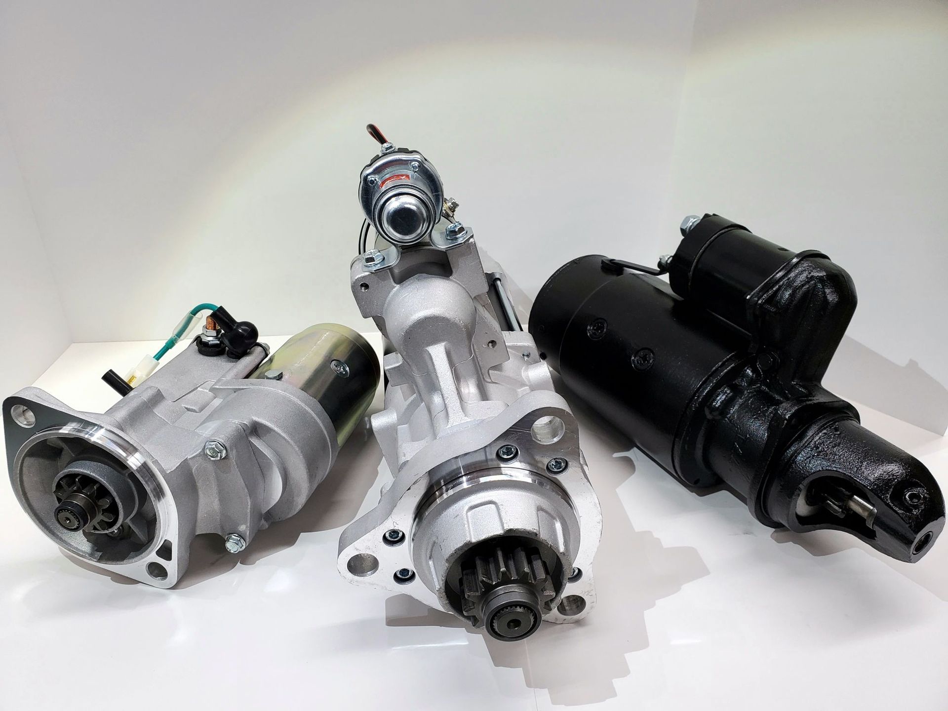 Three metal automotive starter motors arranged on a white background, two silver and one black.