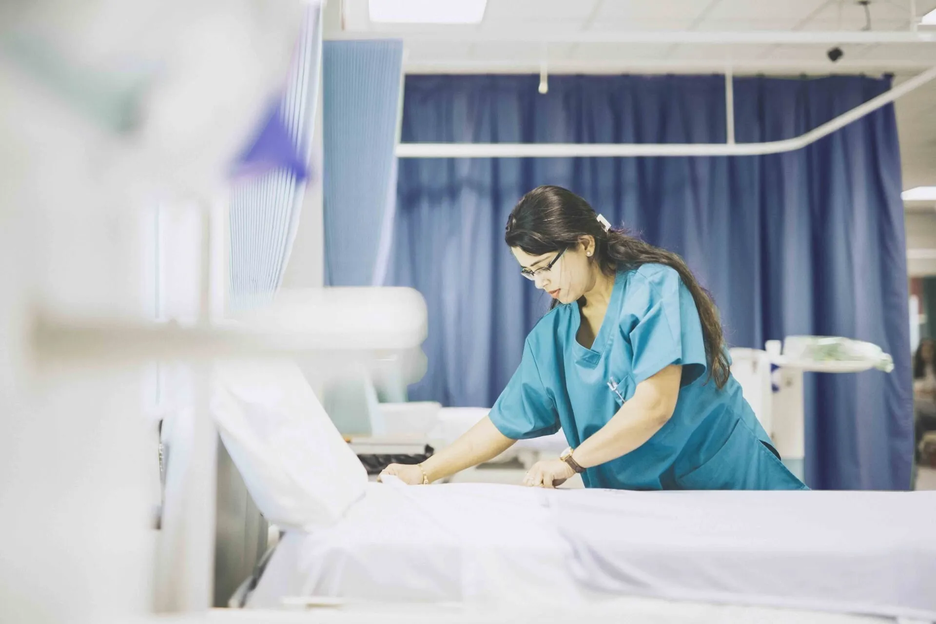 Nurse in blue scrubs making a hospital bed. White sheets and curtains.
