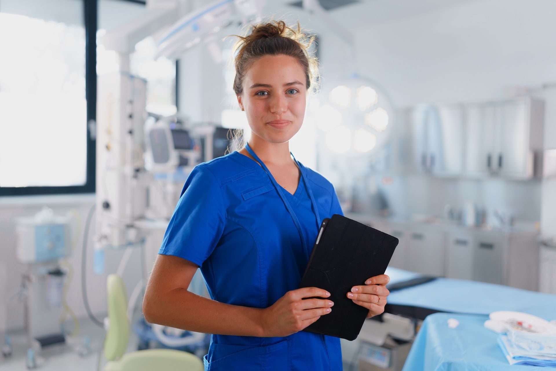 Nurse in blue scrubs holding tablet, smiling in operating room.