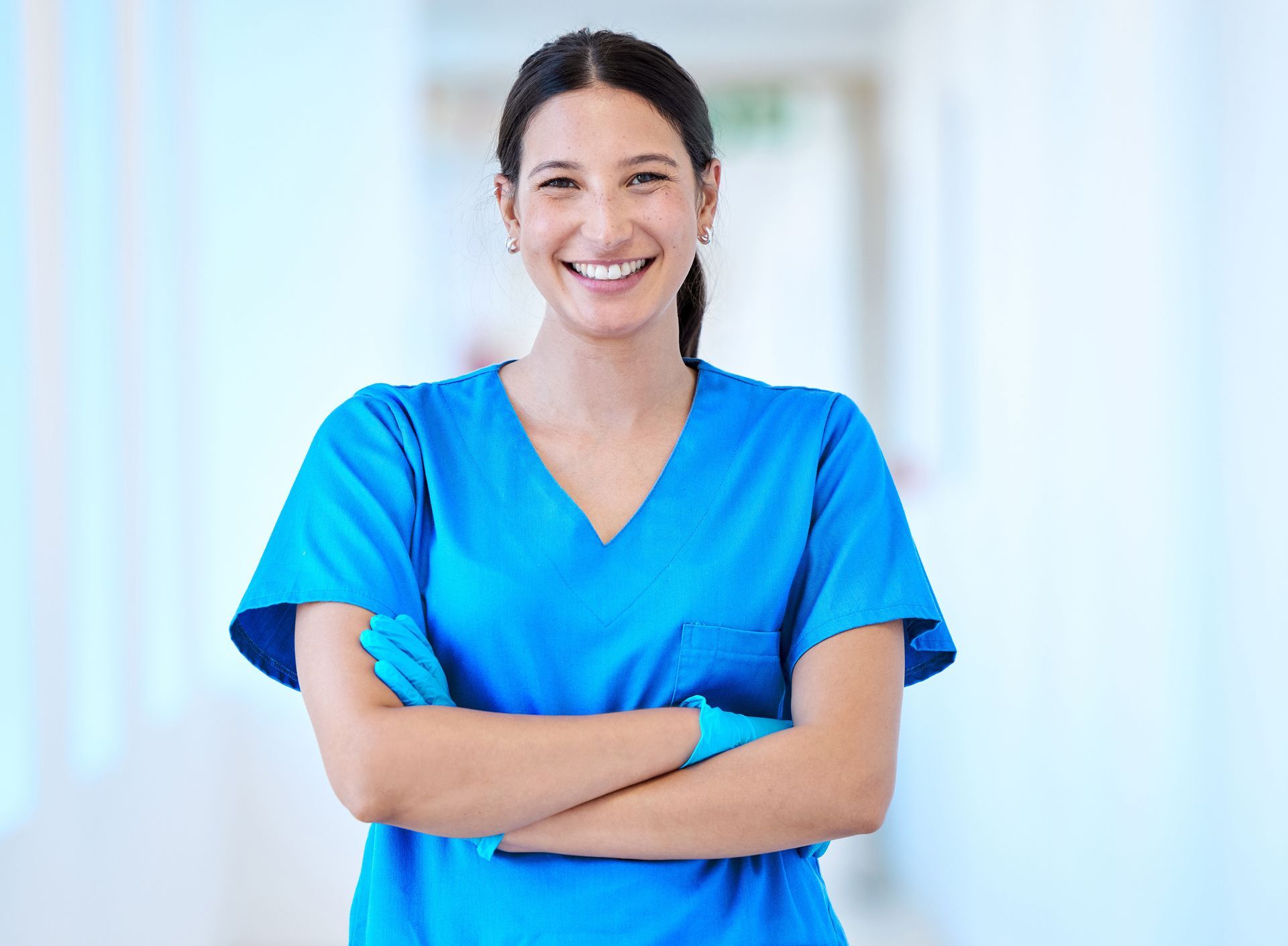 Woman in blue scrubs smiles with arms crossed in a hospital hallway.