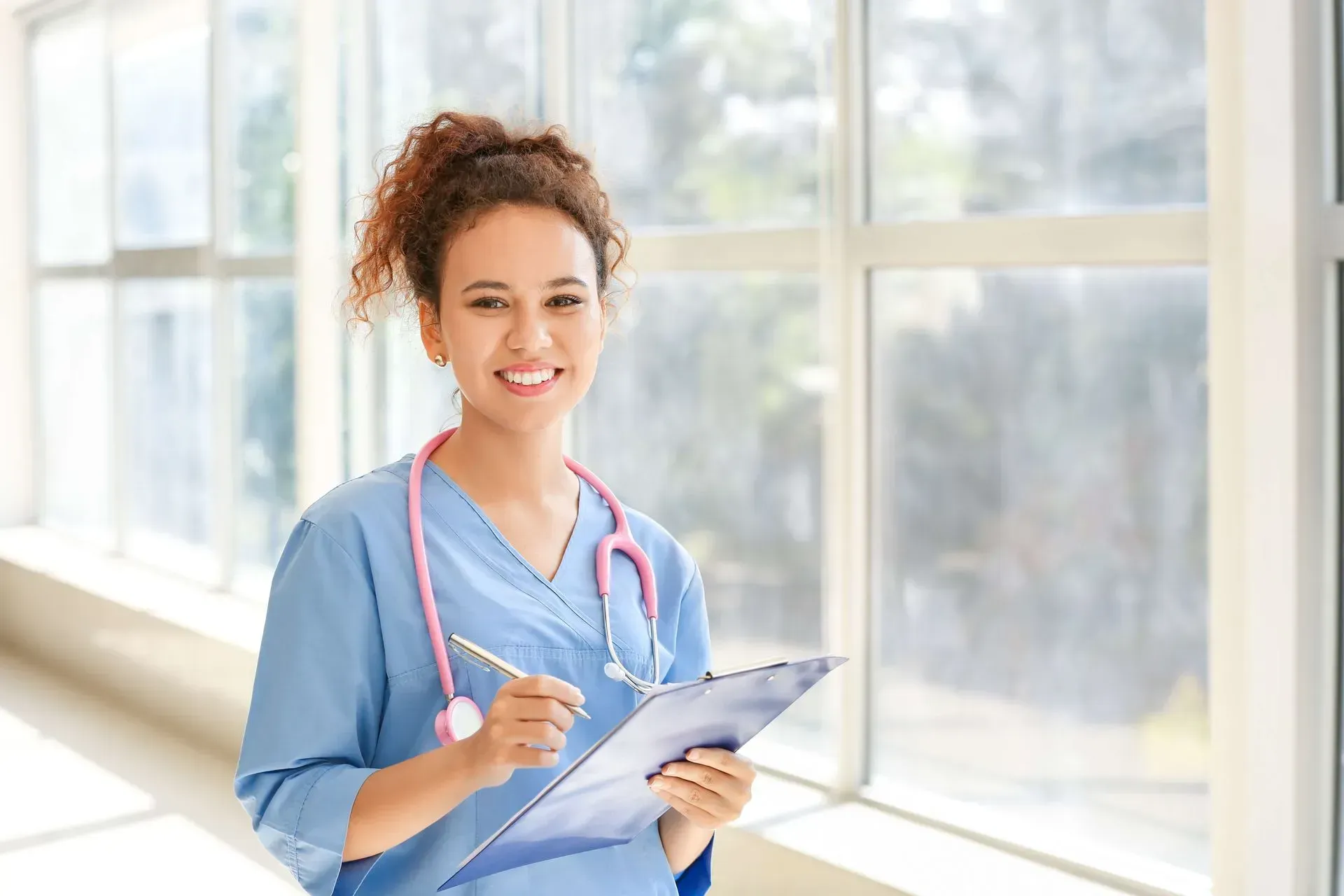 Nurse in blue scrubs holds clipboard and pen, smiles near a window.