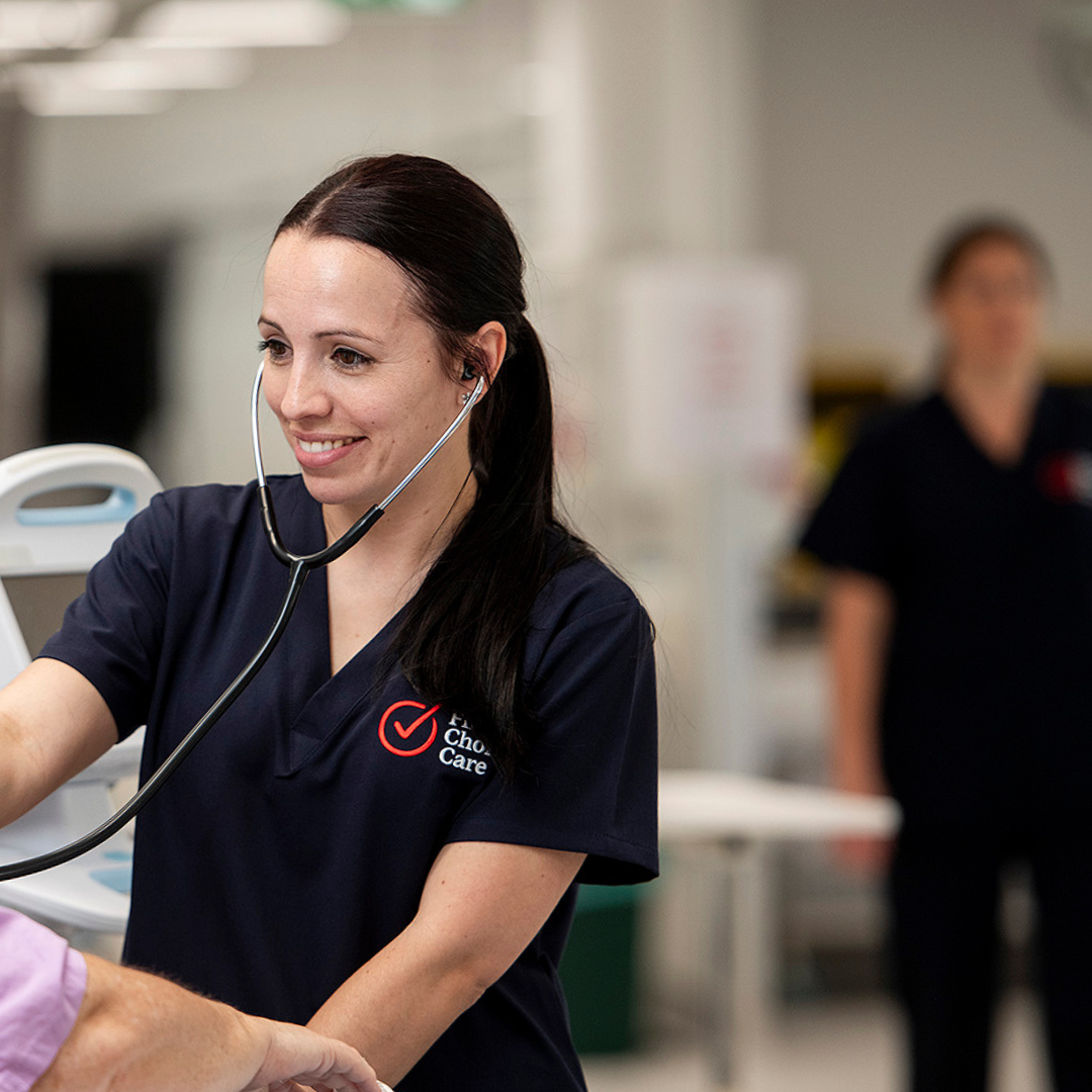 Smiling First Choice Care Nurse in navy scrubs using a stethoscope on a patient in a clinic