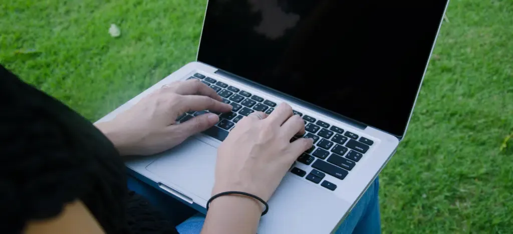 A woman is typing on a laptop computer while sitting in the grass.