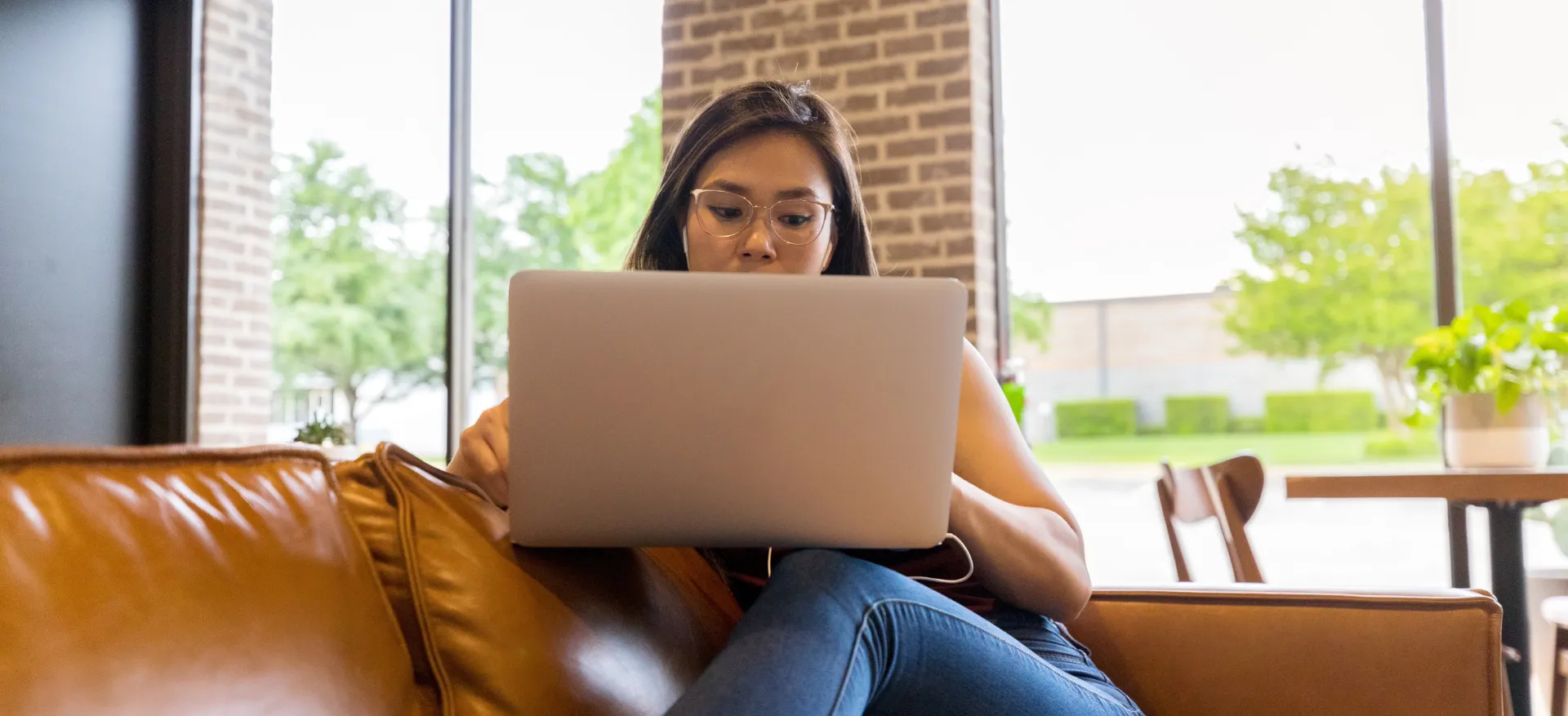 Woman with glasses on laptop, sitting on a brown leather sofa in front of a window.