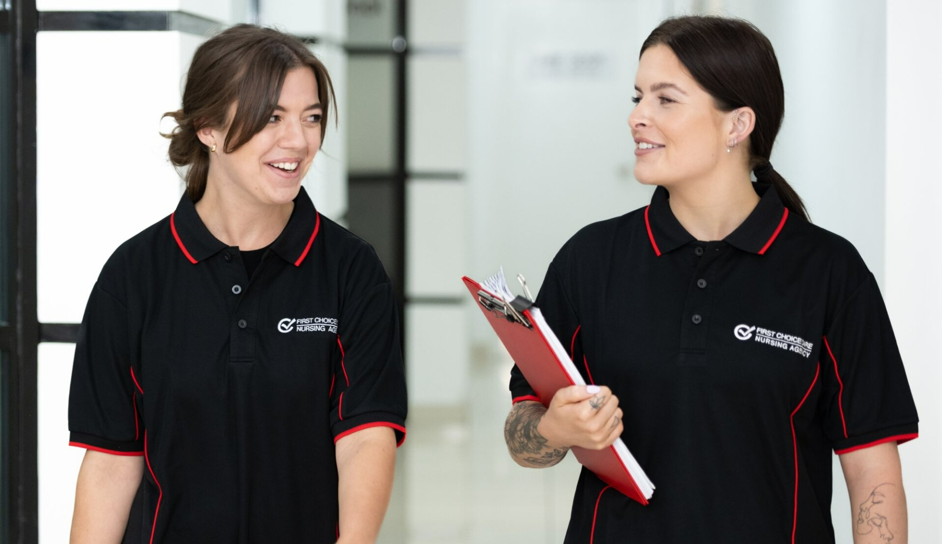 Two women are standing next to each other in a hallway holding a clipboard.