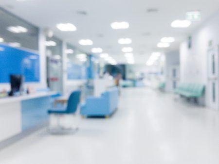 Blurred view of a light-blue hospital hallway with chairs and a reception desk.