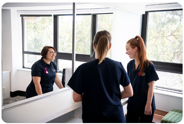 Three women in navy scrubs in a brightly lit room; two face a mirror, one smiles at them.