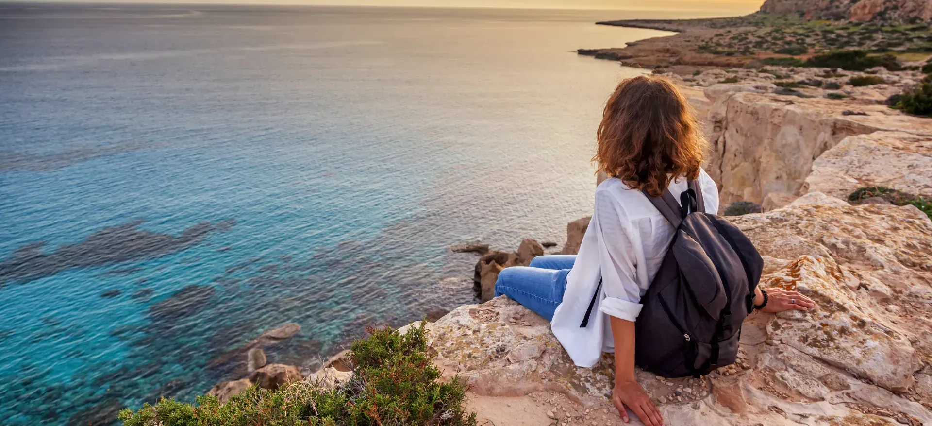 Woman with backpack seated on a cliff, looking at the ocean, sunset.