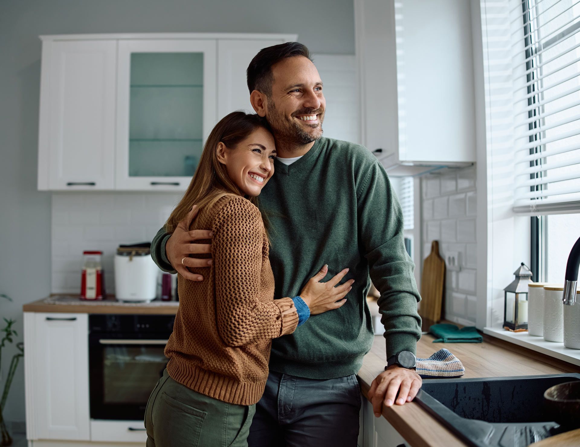 Couple embracing in a kitchen, looking out the window, smiling.