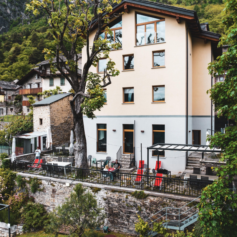 Edificio di tre piani con posti a sedere all'aperto e un grande albero di fronte. Facciata beige con tetto nero in un ambiente montuoso.