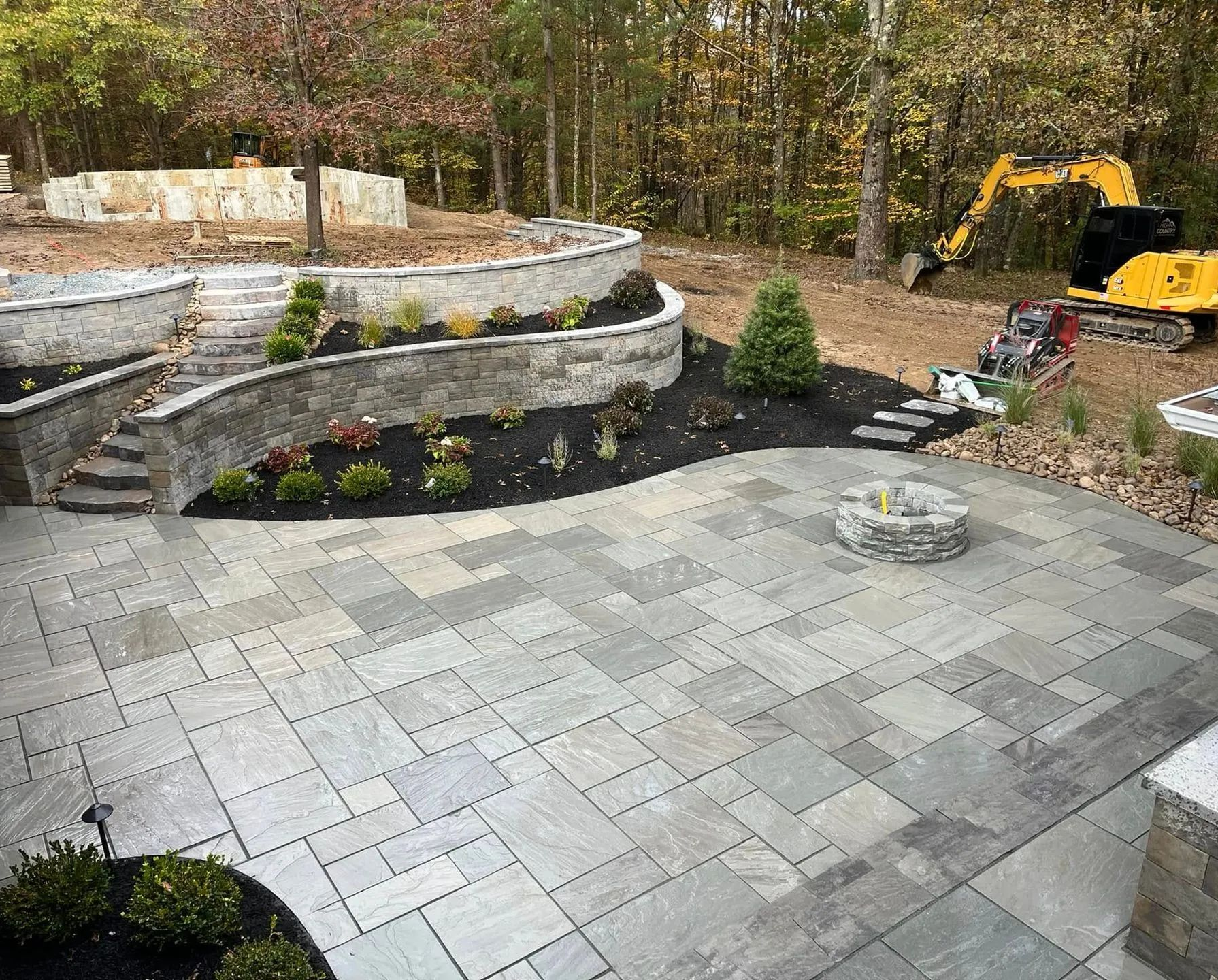 Stone patio with tiered retaining wall, flower beds, and a yellow excavator in a wooded backyard.