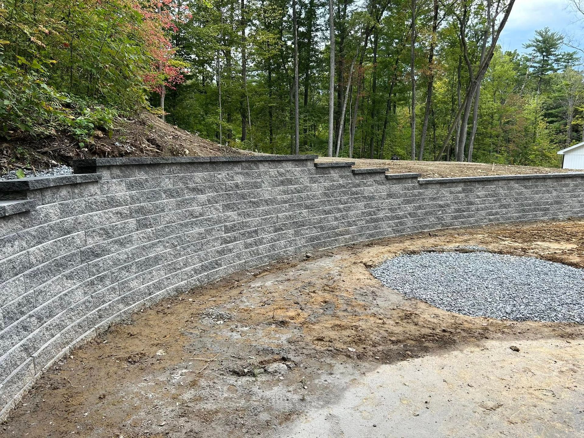 A curved gray stone retaining wall stands in a dirt yard with a circular gravel pit and a forest backdrop.
