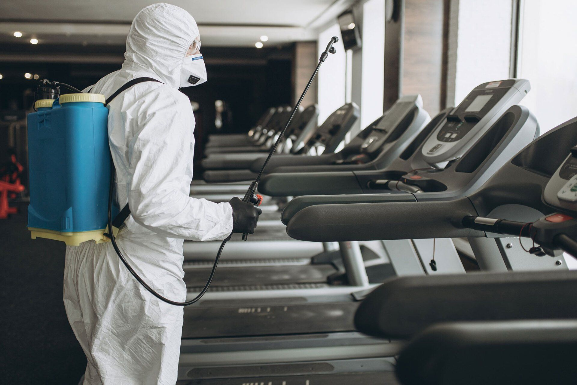 A man in a protective suit is disinfecting treadmills in a gym.