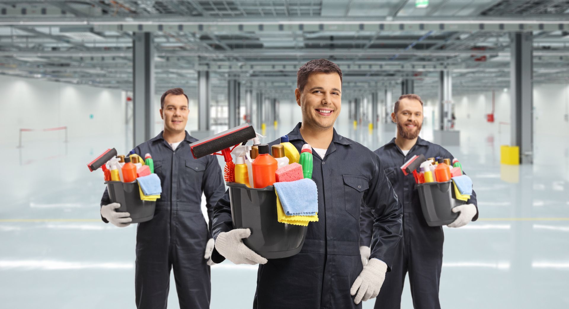 Three men in overalls are holding buckets of cleaning supplies in a warehouse.