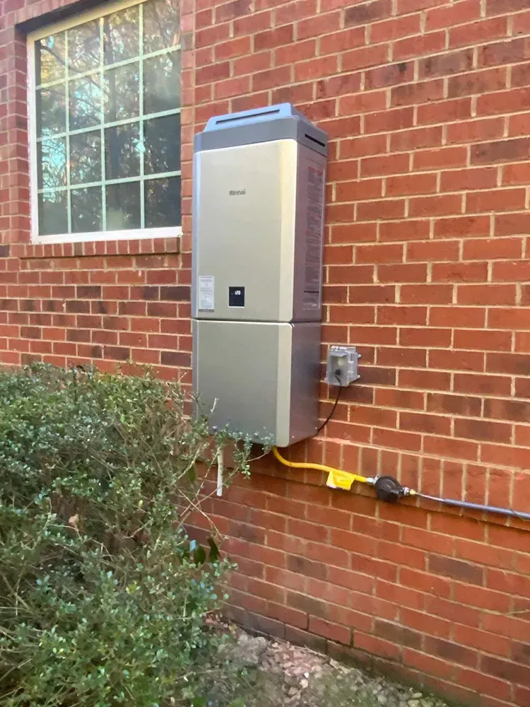 Silver water heater mounted on a red brick wall next to a window and bushes.