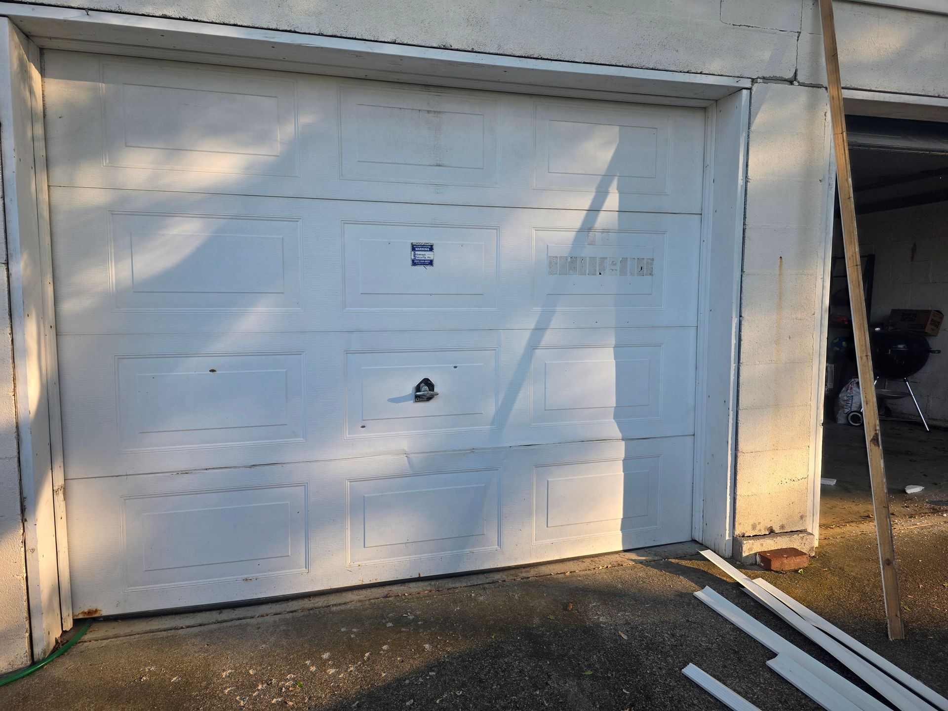 White garage door with a small black handle and sunlight shadows, beside a driveway.