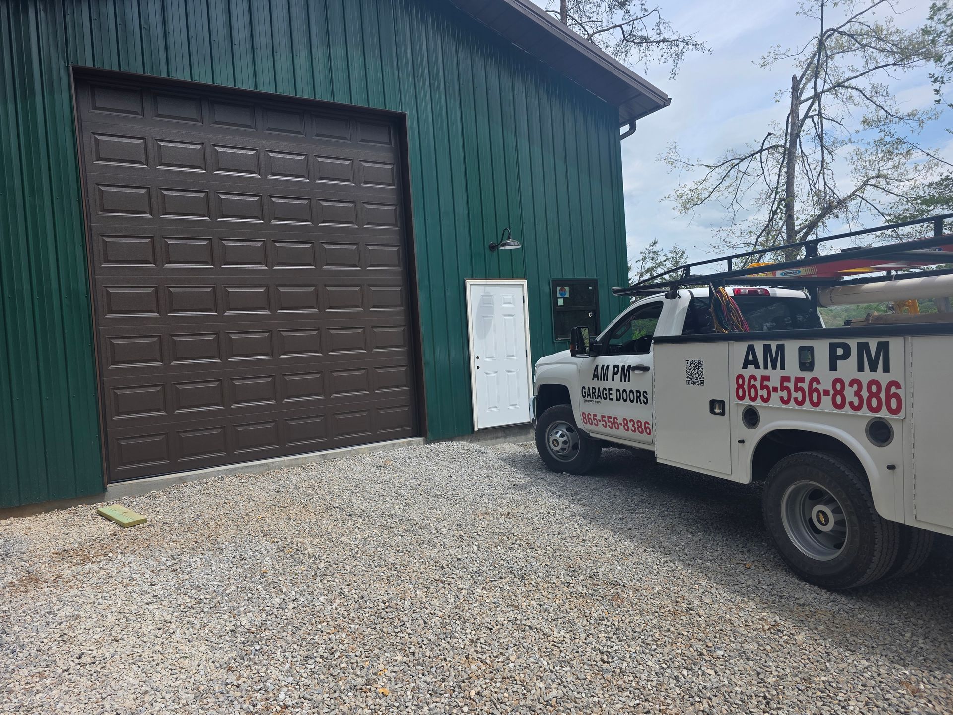 Green building with a large brown garage door and a white service truck parked outside on gravel.