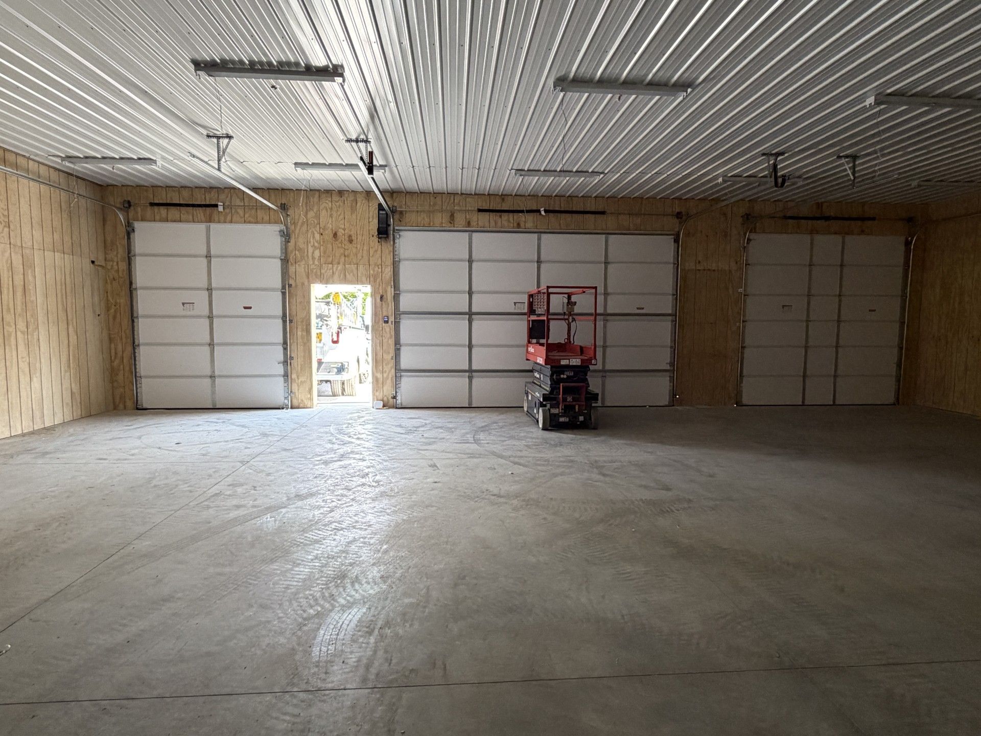 Empty garage with unfinished walls, three large doors, and a red scissor lift near the center.