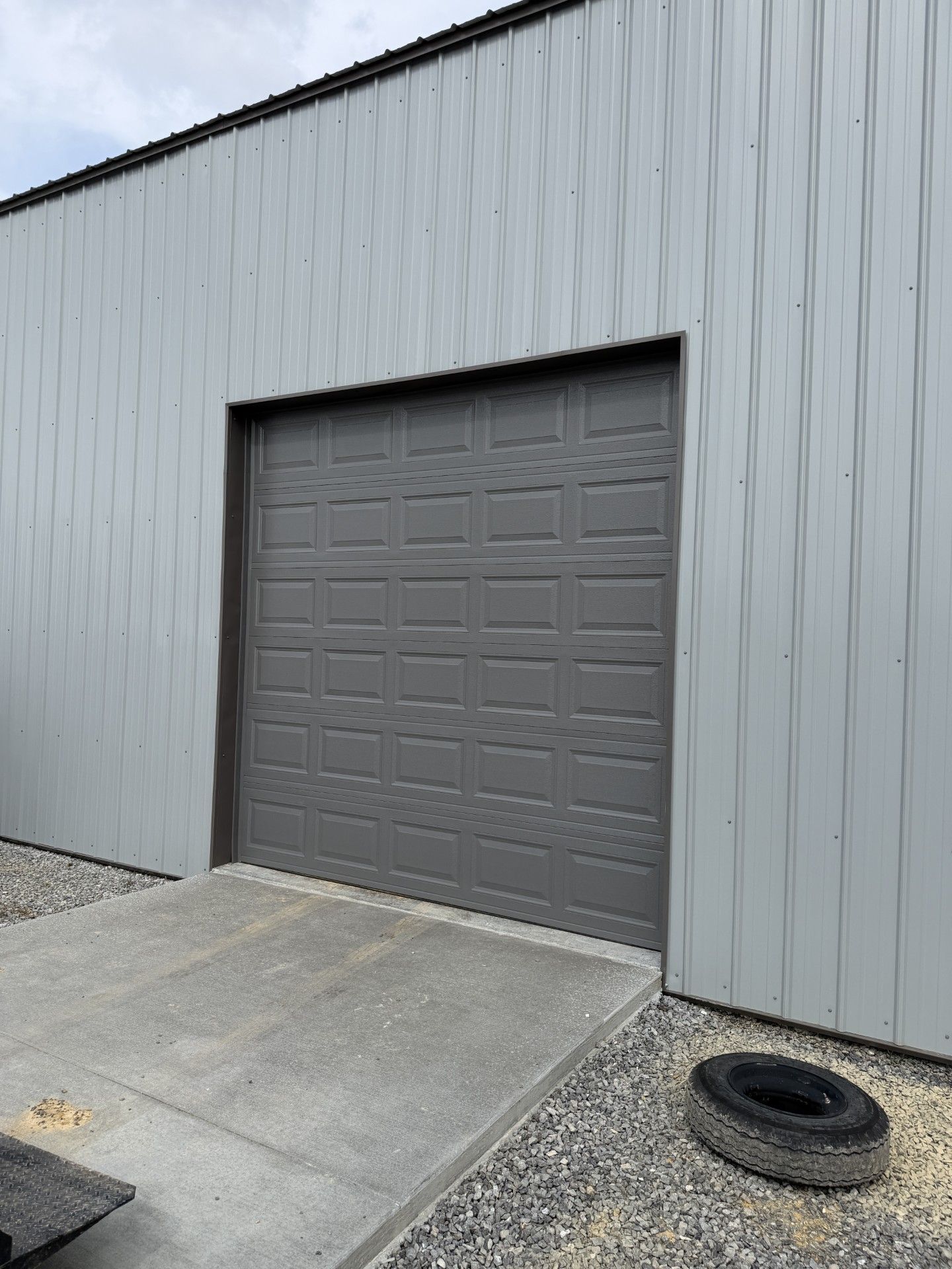 Gray metal garage door on a gray industrial building with a concrete ramp and tire on the ground.