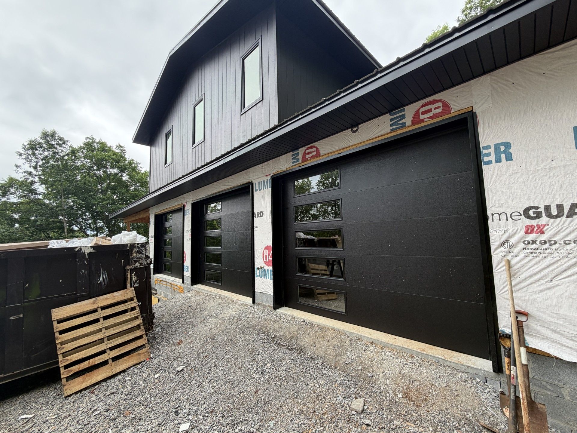 Exterior of a building under construction with black siding, open garage doors, and gravel driveway