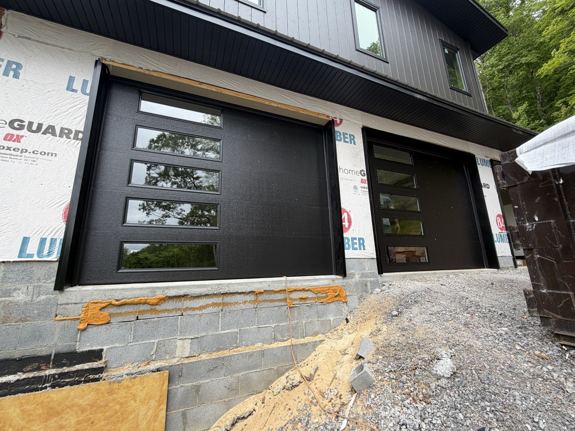 Modern house exterior with black garage doors and unfinished stone foundation beside gravel driveway