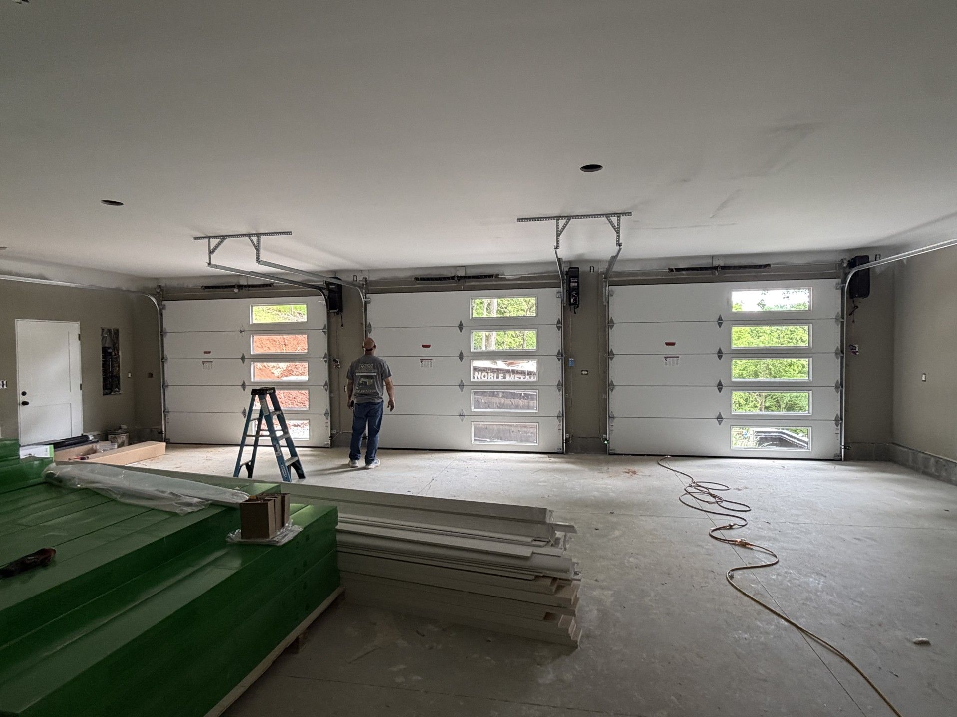 Construction site interior with three garage doors, worker on ladder, and stacked materials on floor